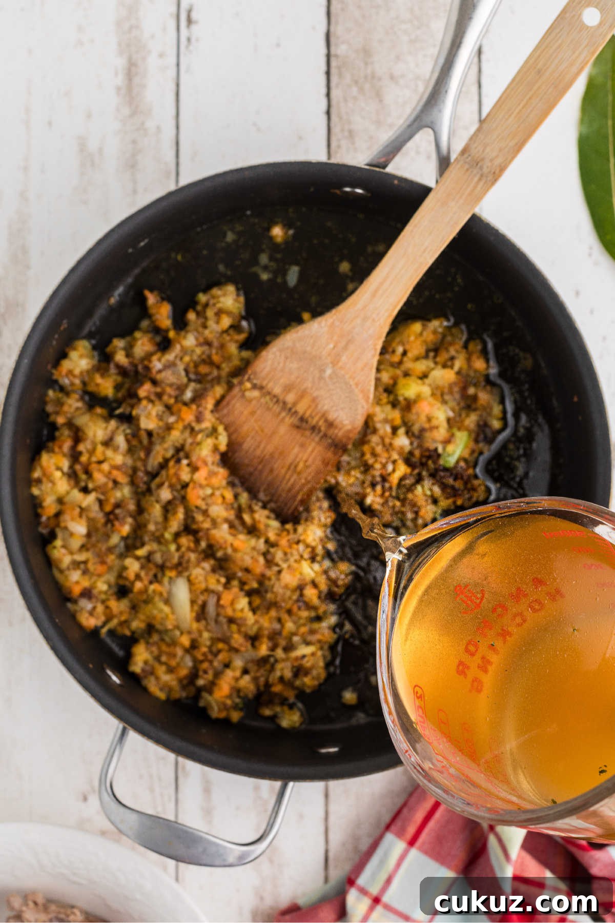 Slowly pouring chicken broth into a large skillet of cooking vegetables.