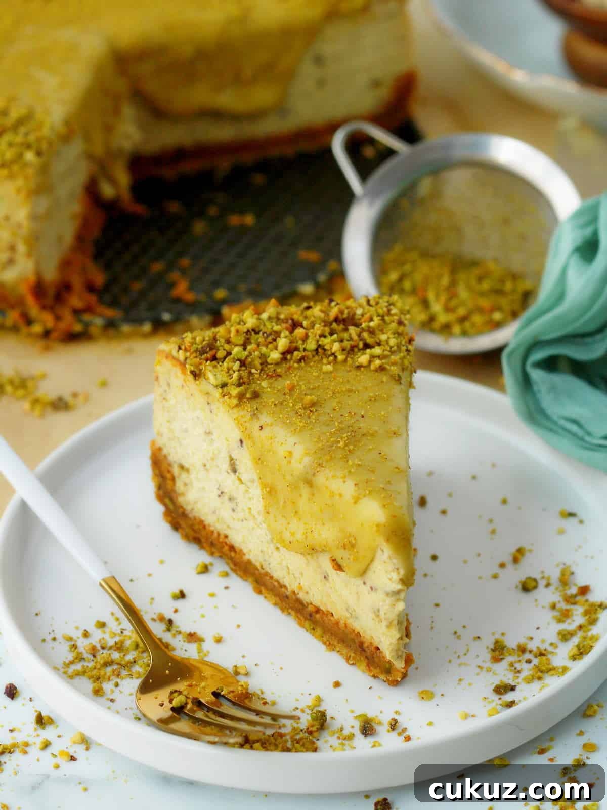 Slice of pistachio cheesecake on white plate, with white fork and whole cheesecake behind it.