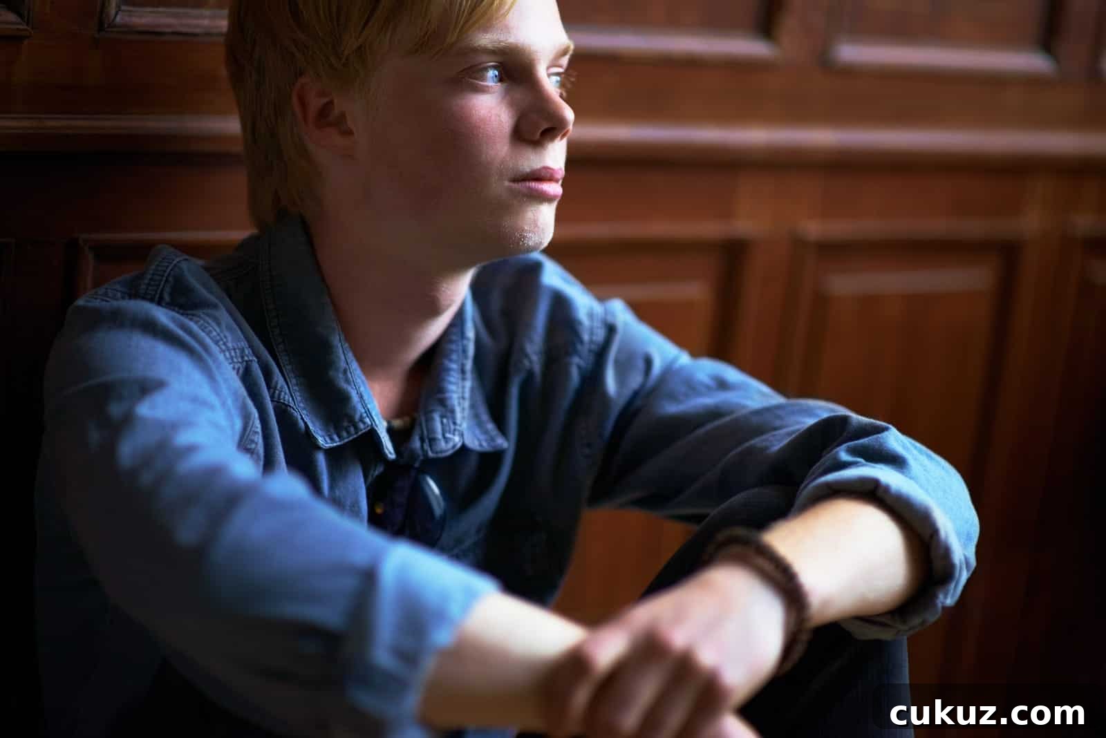 A young person in a denim jacket sits with their arms resting on their knees, gazing thoughtfully to the side. Wood paneling is visible in the background.