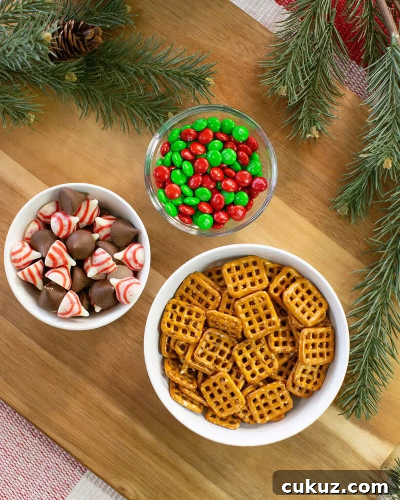 Three bowls on a wooden surface: one with red and green candies, one with square pretzels, and one with chocolate and peppermint candies. Pine branches are placed around the bowls for decoration.