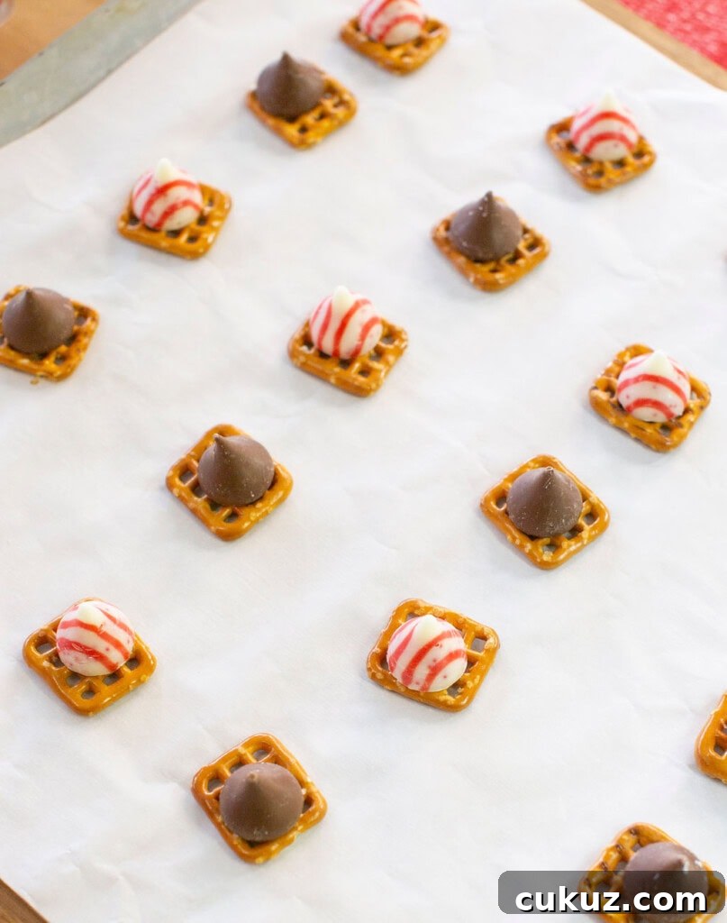Small square pretzels topped with either striped peppermint or chocolate kisses are arranged on a sheet of parchment paper for baking or cooling.