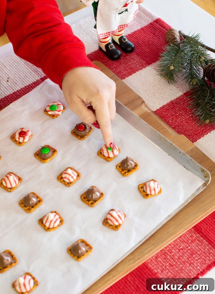 A hand in a red sleeve points to a holiday pretzel treat on a baking sheet lined with parchment paper. The sheet holds various pretzel snacks topped with candy. Festive decor and a red plaid tablecloth are visible nearby.