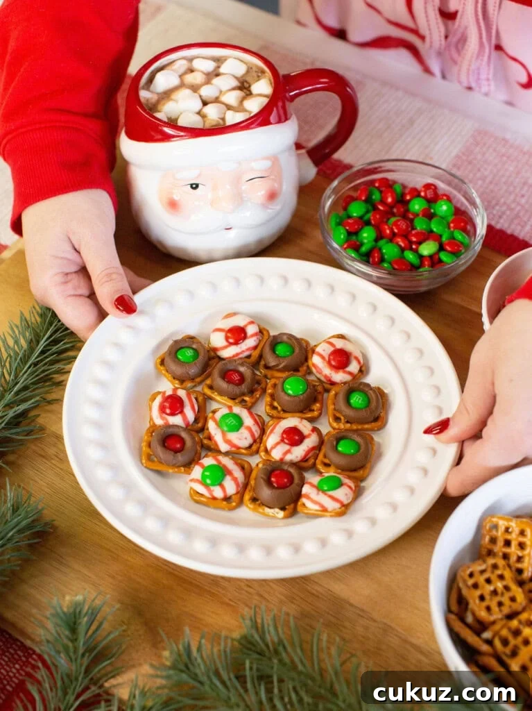 A person in a red sweater holds a plate of holiday pretzel treats topped with chocolate, candy, and festive colors. Nearby are a Santa mug with hot chocolate and marshmallows, a bowl of red and green candies, and some pine branches.
