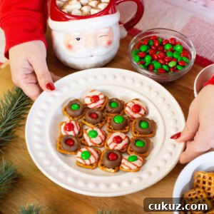 A person in a red sweater holds a plate of holiday pretzel treats topped with chocolate, candy, and festive colors. Nearby are a Santa mug with hot chocolate and marshmallows, a bowl of red and green candies, and some pine branches.