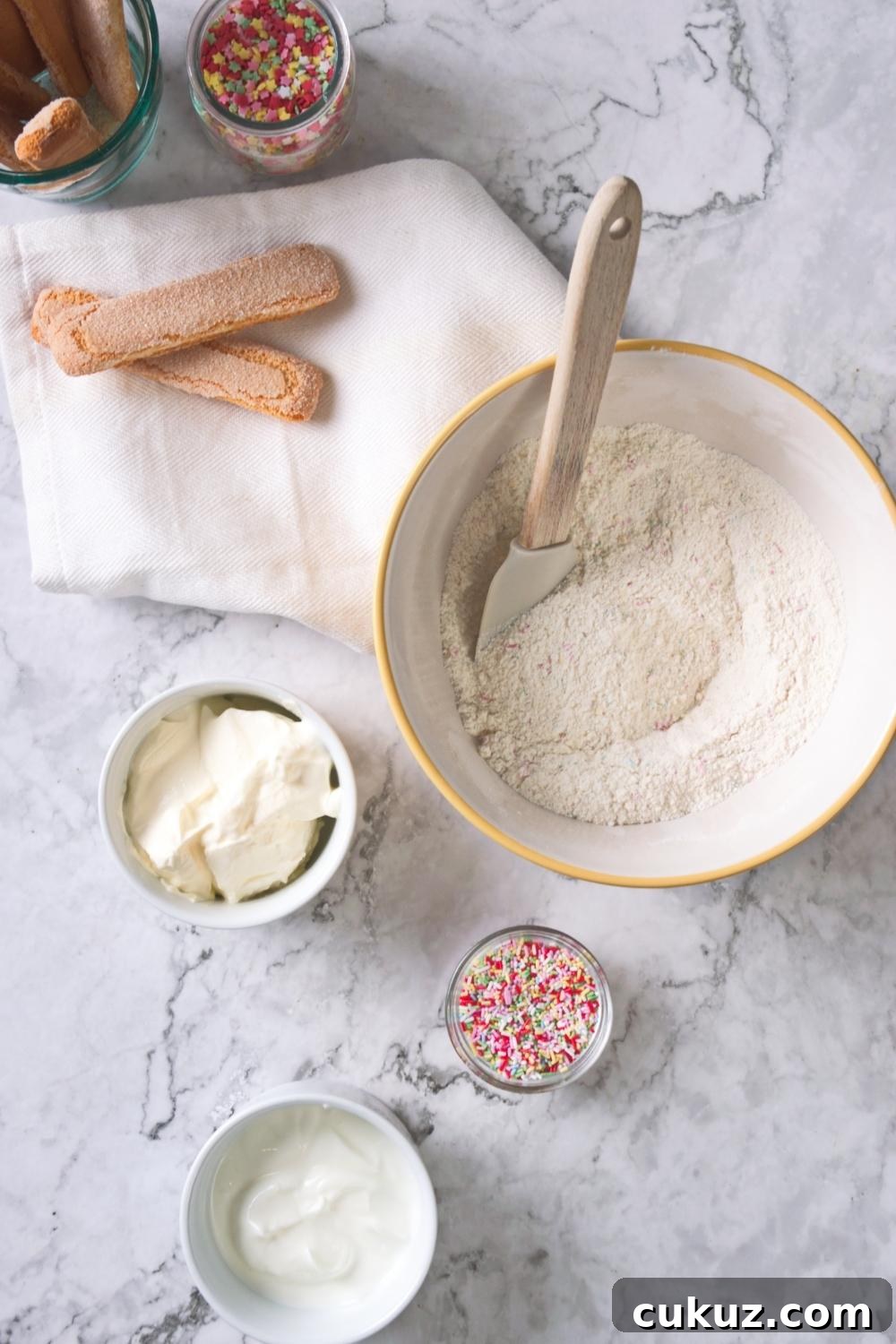 Quick 4-Ingredient Dunkaroo Dip Ready in 5 Minutes 4 An overhead view of a large mixing bowl containing funfetti cake mix, vanilla yogurt, Cool Whip, and a small bowl containing rainbow sprinkles.