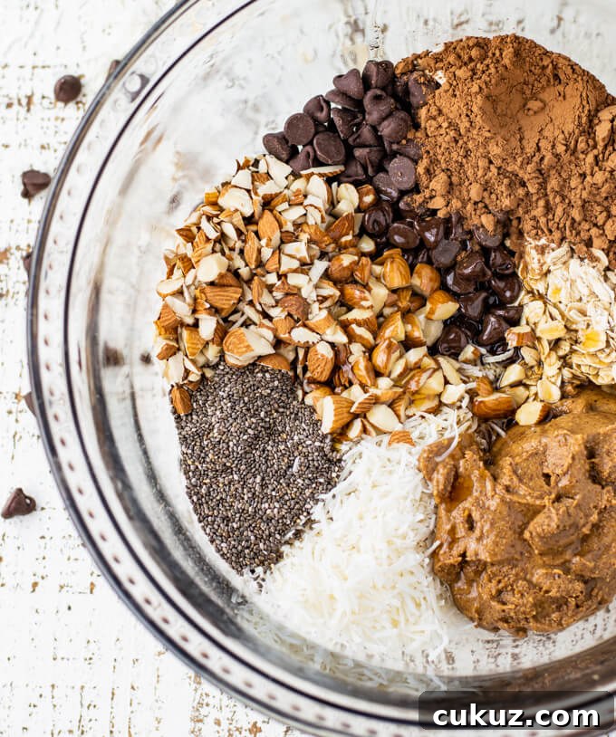 A clear mixing bowl of chopped almonds, chocolate chips, cocoa, almond butter, oats, coconut flakes and chia seeds.