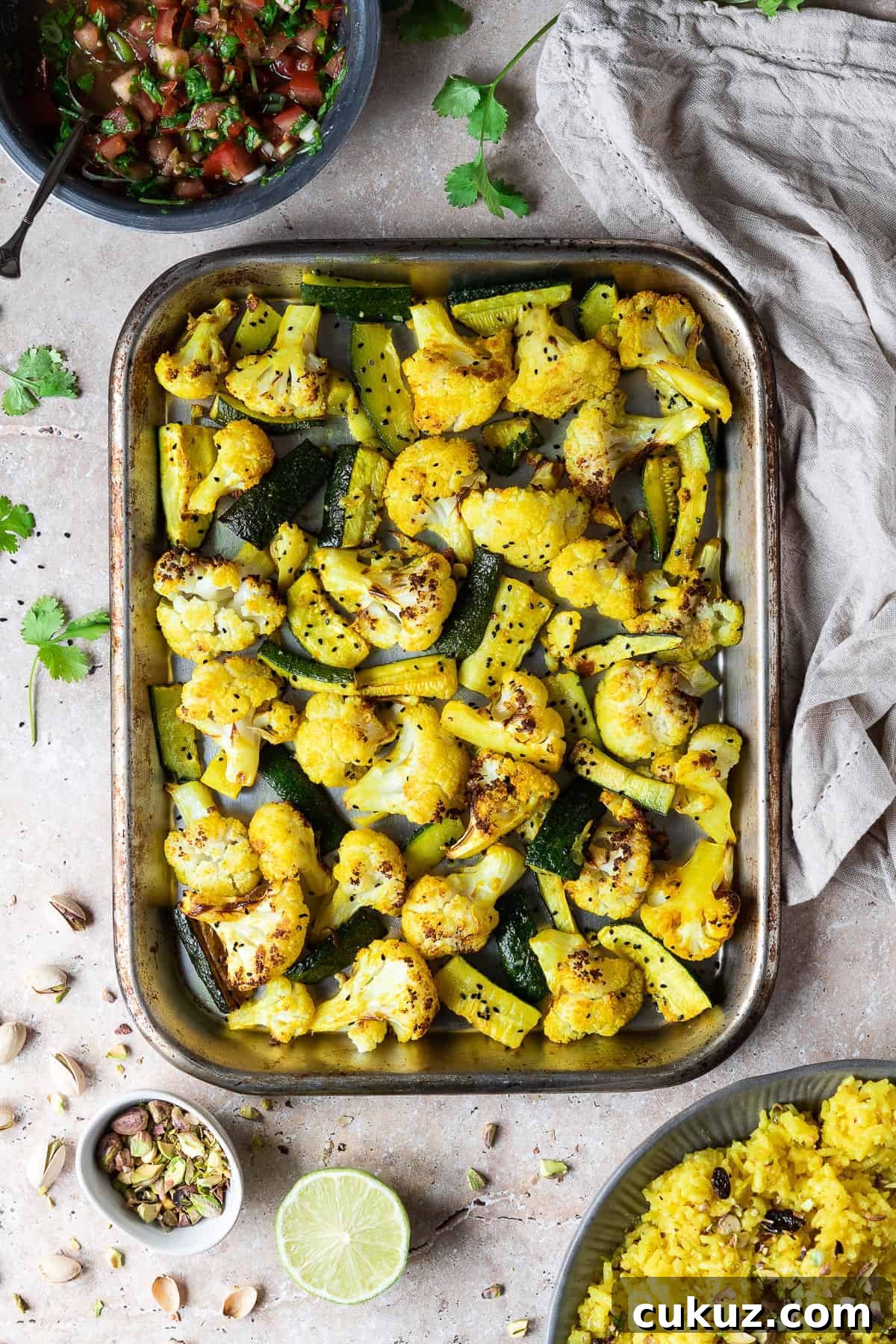 A roasting tray of spiced cauliflower and courgette with a bowl of yellow rice, a bowl of tomato salsa, a cloth, half a lime, pistachios and fresh coriander leaves.