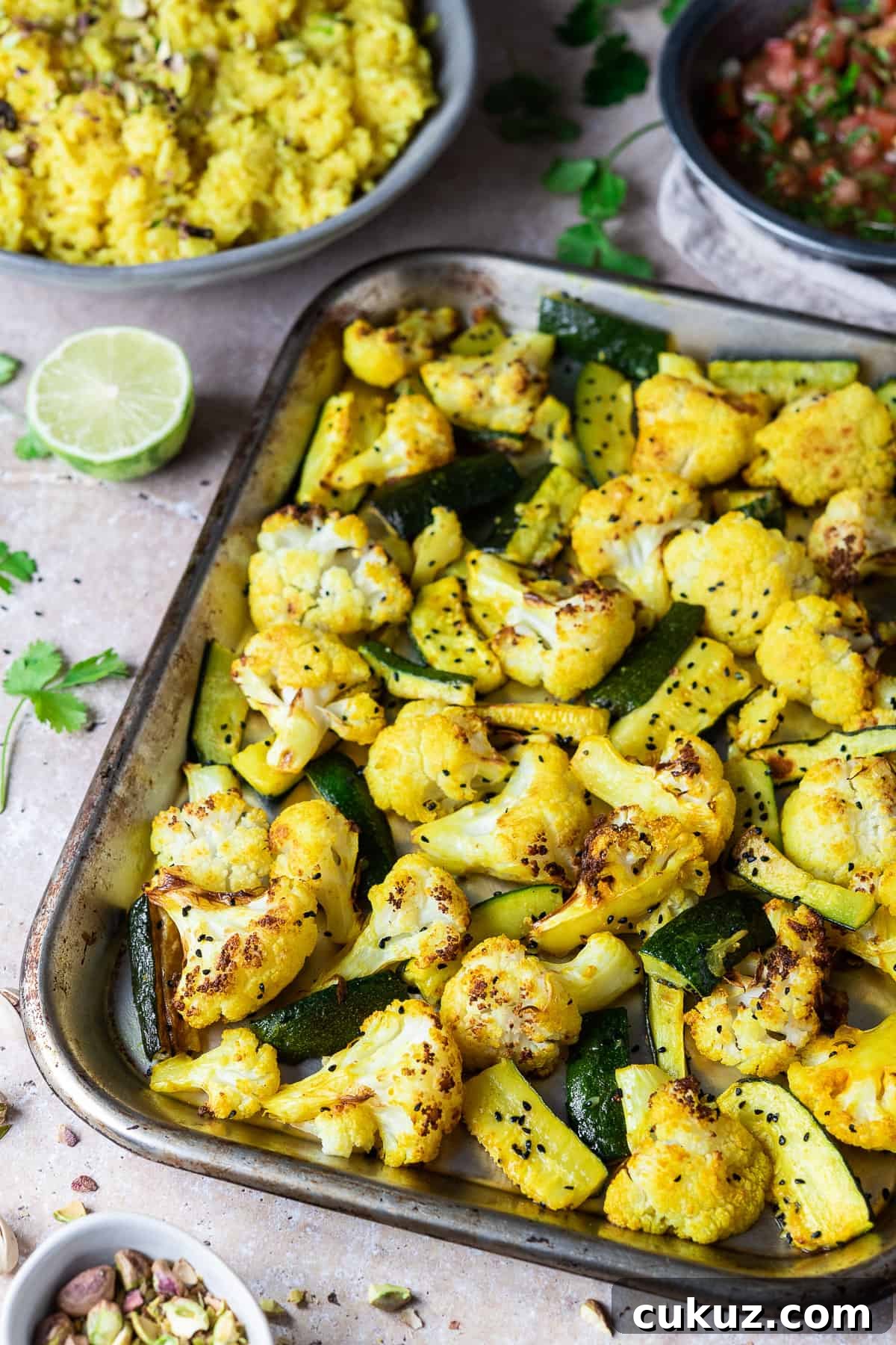 A tray of spiced roasted cauliflower and courgette on a stone bakground with a bowl of yellow rice and a bowl of salsa.