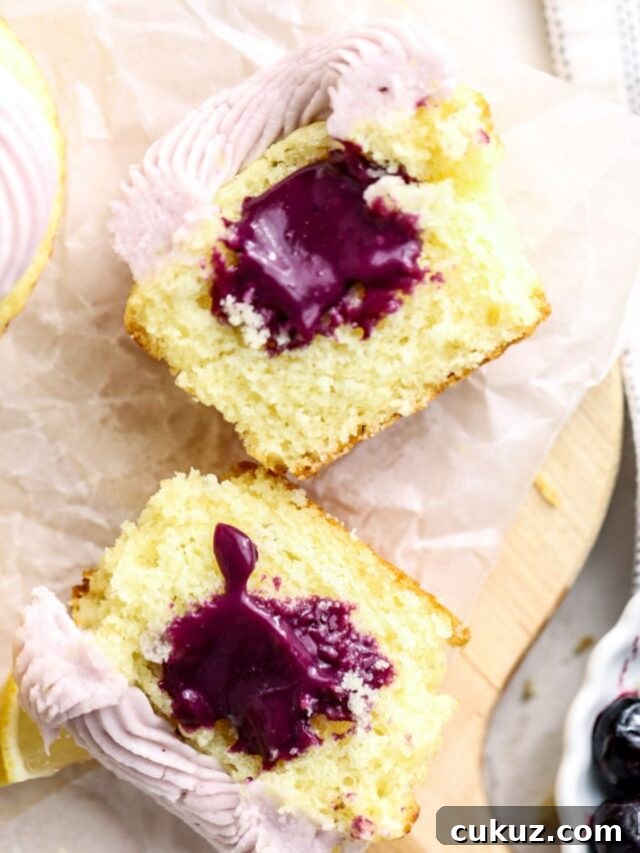A frosted cupcake cut in half with a purple blueberry filling and a bowl of blueberries.