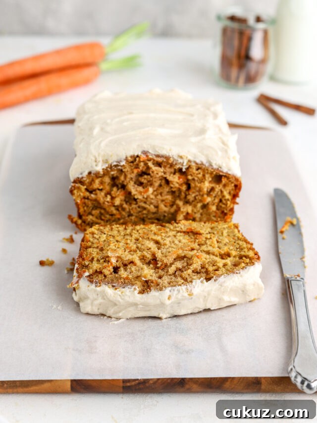 A loaf cake with frosting, a knife, and some carrots and cinnamon sticks.