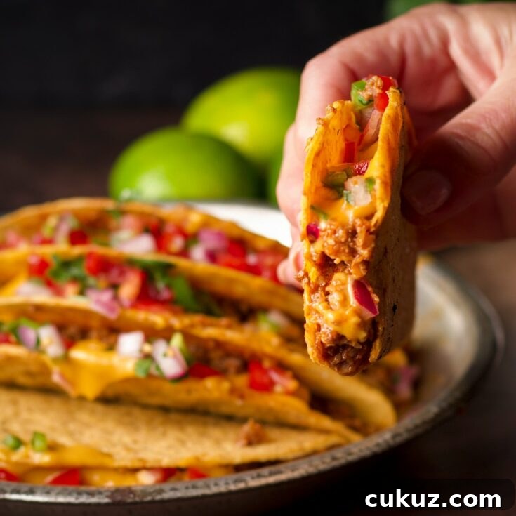 Someone lifting a hard shell taco filled with vegan taco meat and vegan nacho cheese sauce from a plate of tacos.