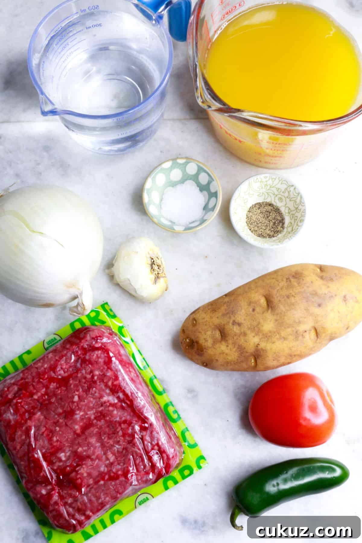 Measuring cup with water, another with chicken broth, an onion, a small bowl with salt, another with pepper, a garlic bulb, a pound of ground beef, a potato, a tomato, and a jalapeño