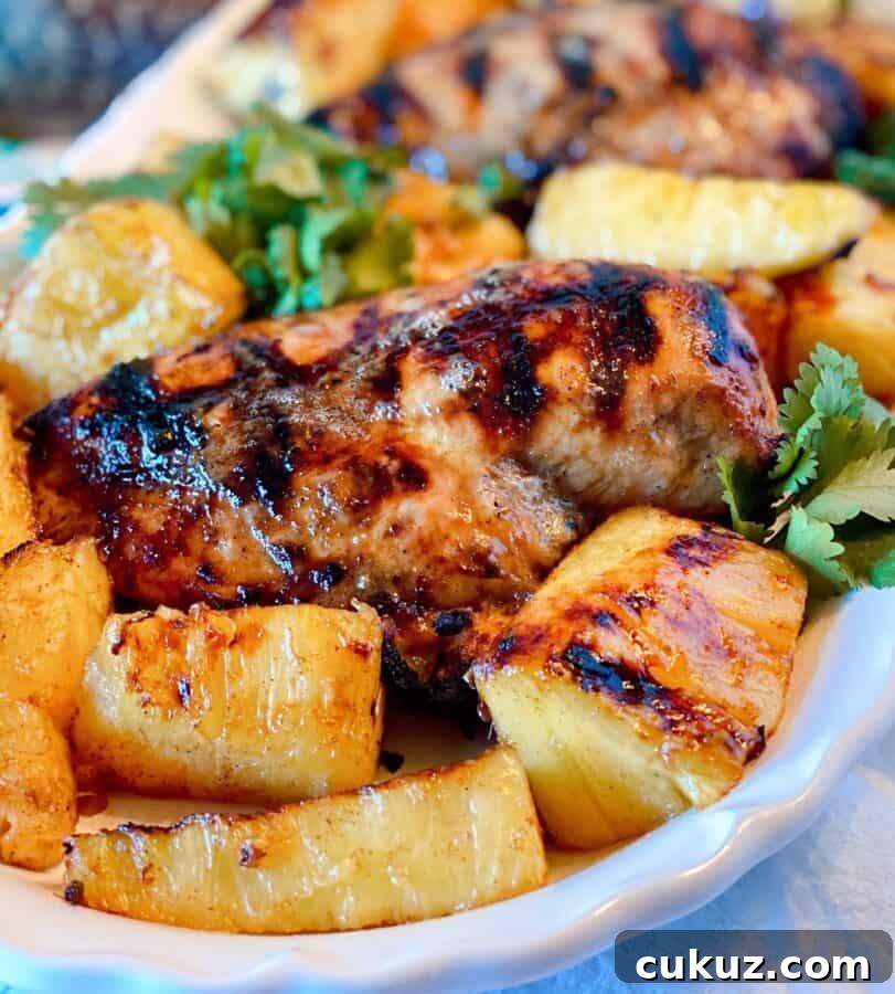 Close-up photo of a grilled jerk chicken breast on a white platter with grilled pineapple spears.