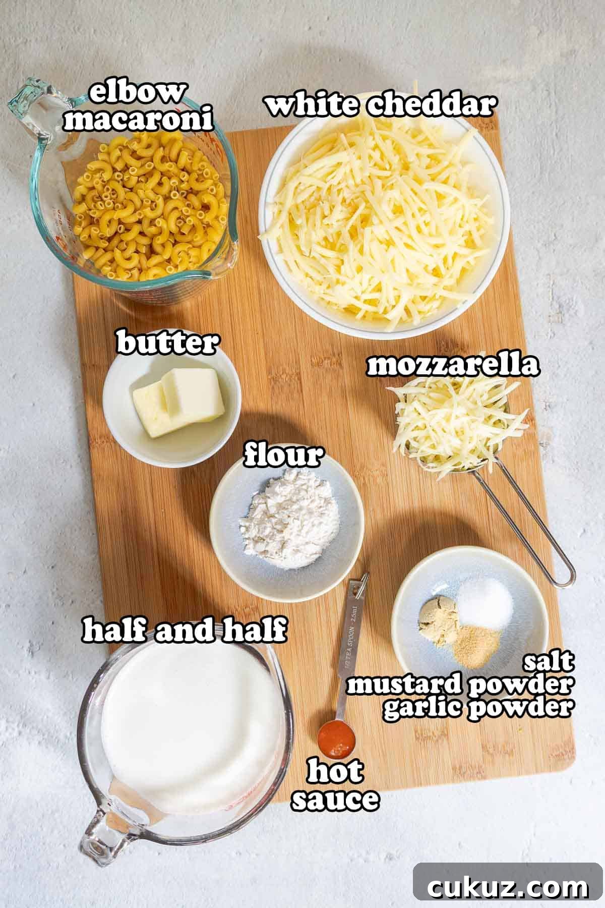Ingredients for white cheddar mac and cheese arranged on a board. Text on the image says 'elbow macaroni', 'white cheddar', 'butter', 'mozzarella', 'flour', 'half and half', 'hot sauce', 'salt', 'mustard powder', 'garlic powder'.