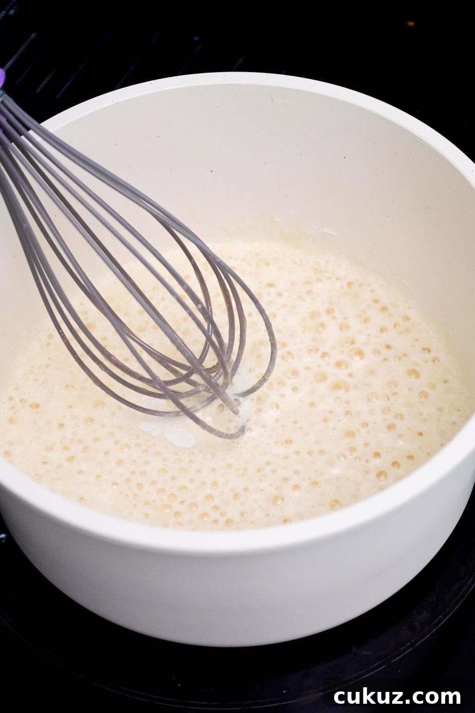 A whisk stirring bubbling melted butter and flour in a white pot.