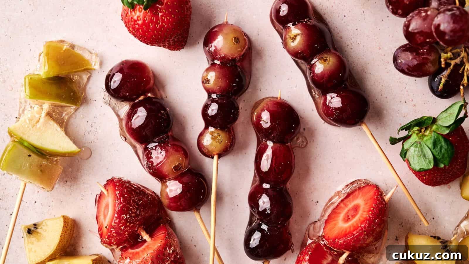 Skewers of grapes and strawberries coated in a shiny glaze are arranged on a light surface with loose fruit pieces nearby.