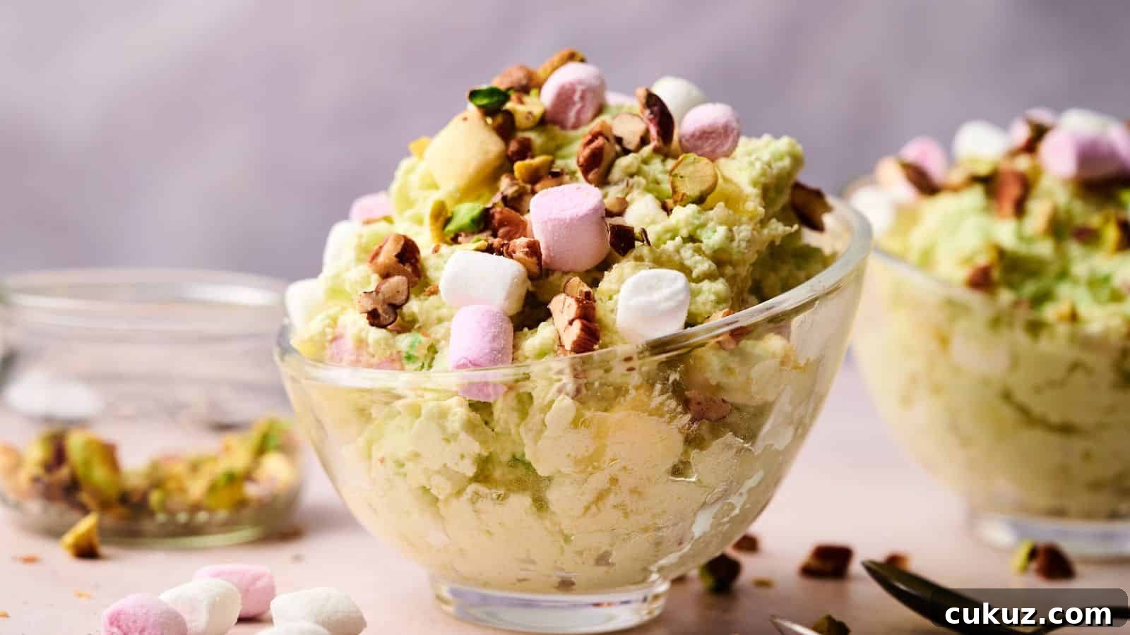 A glass bowl filled with pistachio-flavored dessert topped with mini marshmallows and chopped nuts, with another similar bowl in the background.