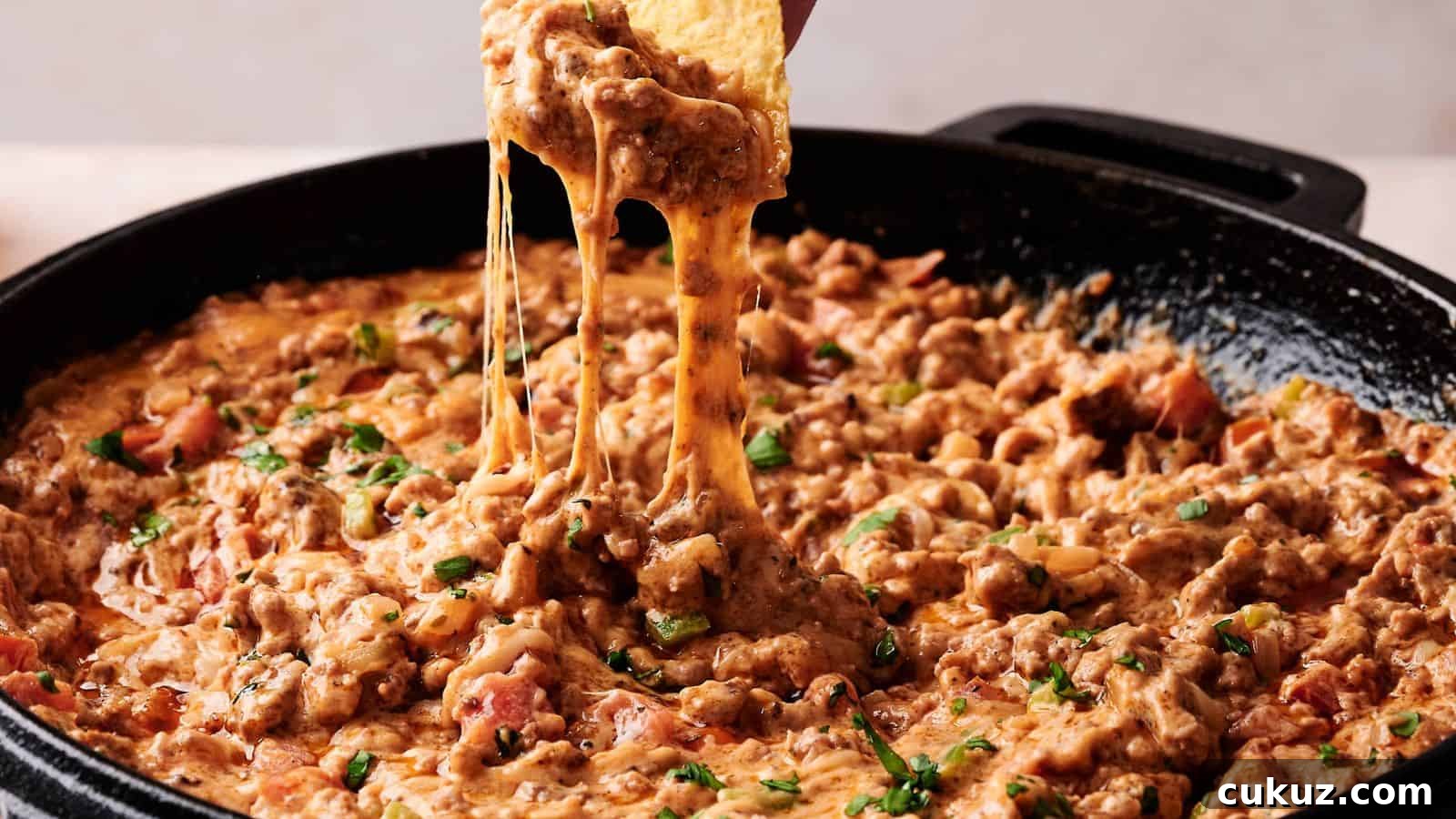 A tortilla chip is dipped into a skillet of cheesy ground beef and tomato dip, with melted cheese stretching between the chip and the dip.