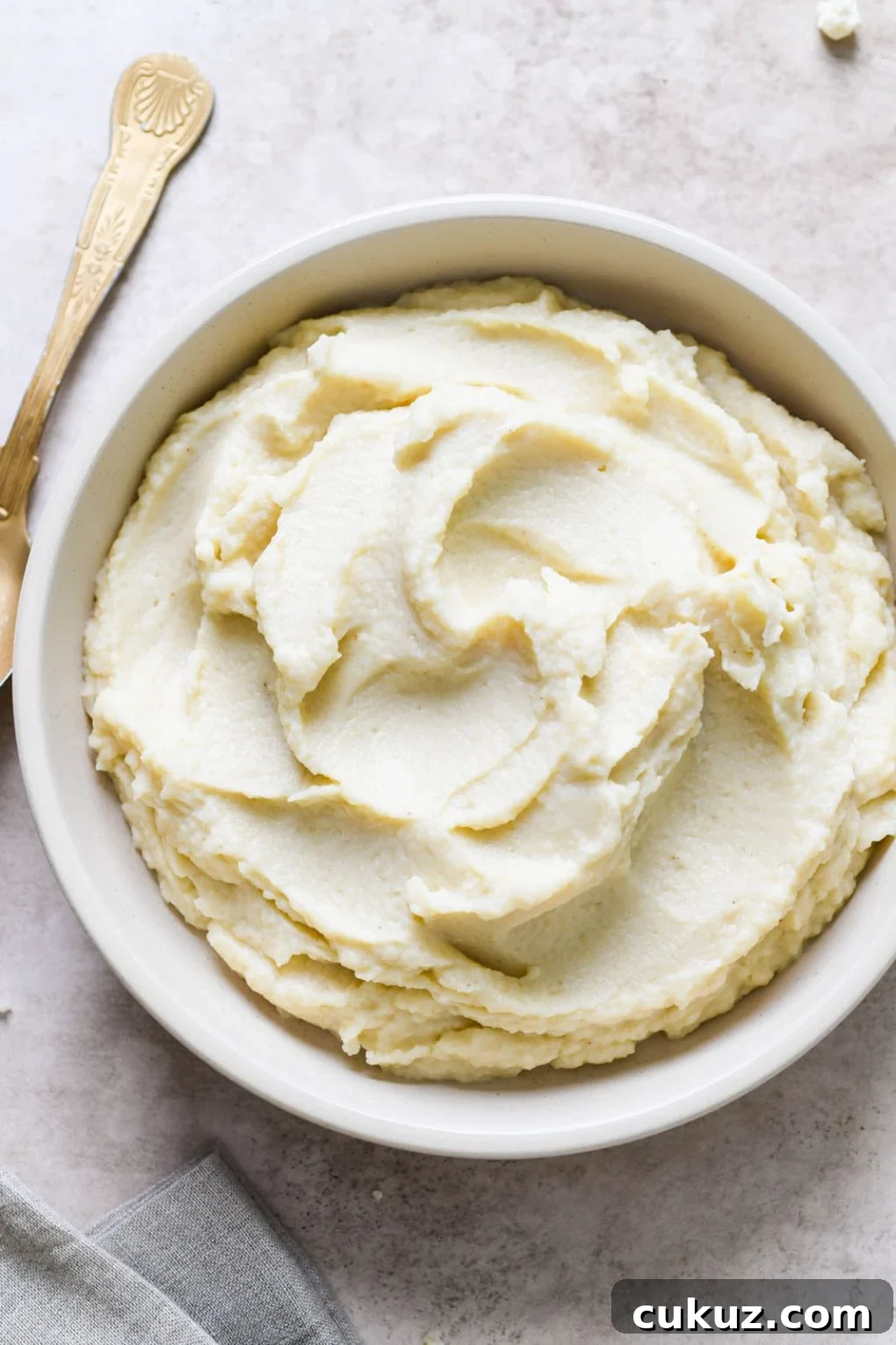 Overhead close up view of swooping mashed cauliflower in a round white bowl, next to a gold antique spoon, on a light grey background. 