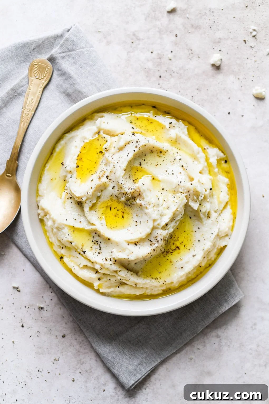 Overhead shot of a large white bowl filled with swooping cauliflower mash, drizzled with bright yellow melted ghee, and sprinkled with cracked black pepper. On a light grey background, next to a slanted antique gold spoon. 
