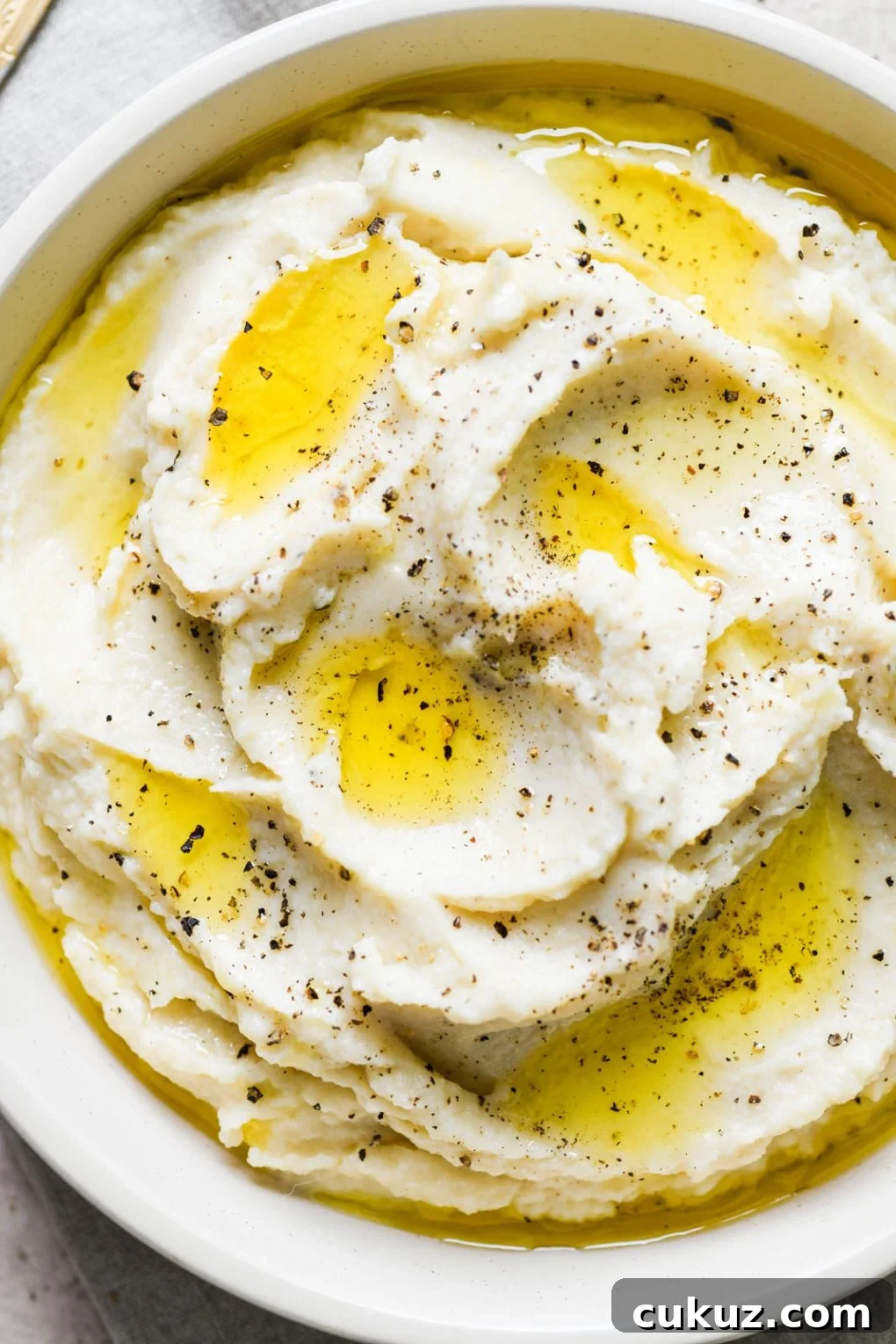 Super close up macro shot of a large white bowl filled with swooping cauliflower mash, drizzled with bright yellow melted ghee, and sprinkled with cracked black pepper. 