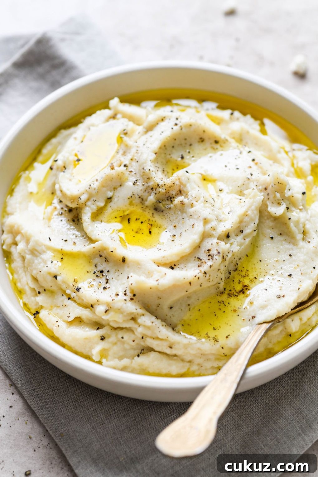 45 degree angle shot of a large white bowl filled with swooping cauliflower mash, drizzled with bright yellow melted ghee, and sprinkled with cracked black pepper. On a light grey background, with a gold antique spoon dipped into the bowl on the side. 