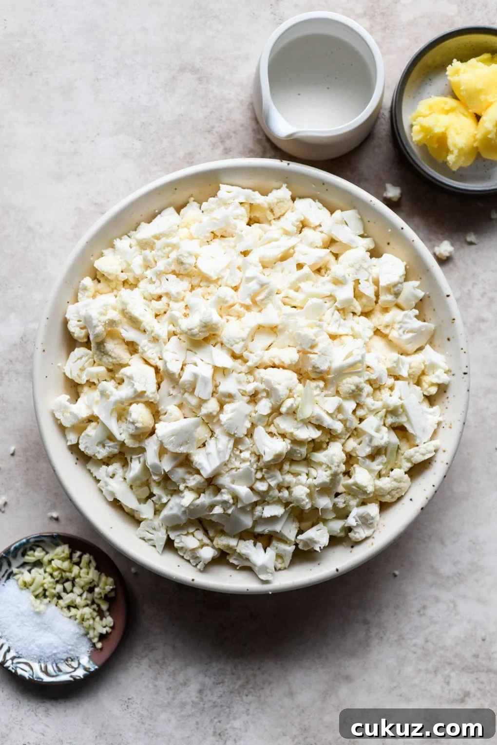 Ingredients for the creamiest Whole30 mashed cauliflower. A large white bowl in the center filled with chopped raw cauliflower, next to a small dish with chopped garlic and sea salt, a dish with 3 tablespoons of ghee, and a small ceramic pitcher with non dairy milk. On a light grey background. 