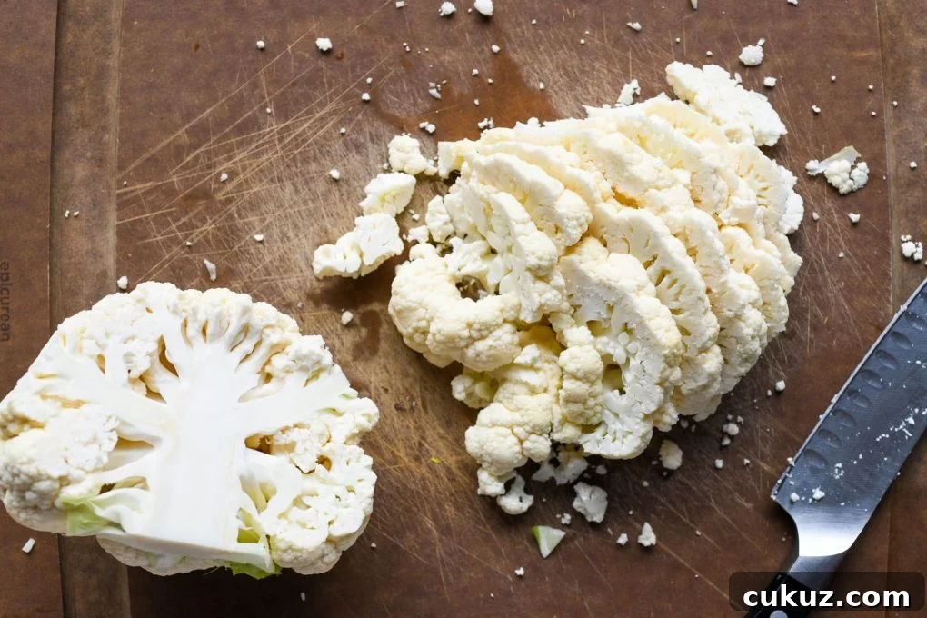 Overhead shot of a cauliflower on a dark brown cauliflower. Cauliflower is cut in half, one half is set aside, the other has been sliced into thin slices.