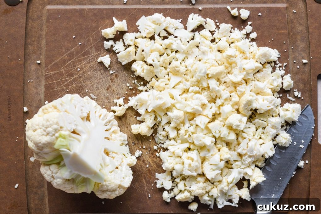 Overhead shot of a cauliflower on a dark brown cauliflower. Cauliflower is cut in half, one half is set aside, the other has been chopped up into super small pieces.