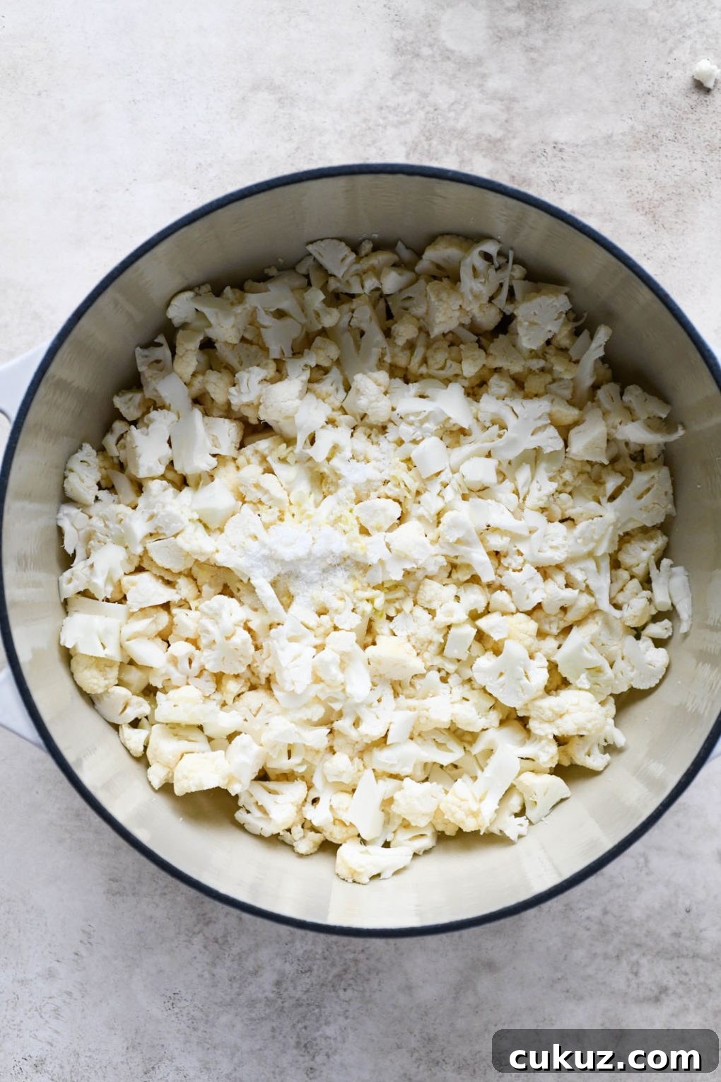 Overhead shot of raw cauliflower inside large white ceramic dutch oven, on a light grey background. 