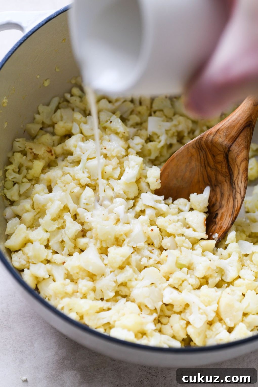 Overhead shot of cooked cauliflower inside large white ceramic dutch oven, on a light grey background. A hand is pouring non dairy milk into the pot with the cooked cauliflower. 