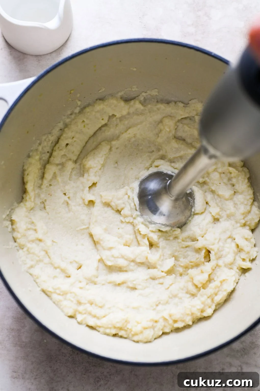 Overhead picture of a handheld immersion blender processing the cooked cauliflower inside white ceramic dutch oven. On a light grey background. 