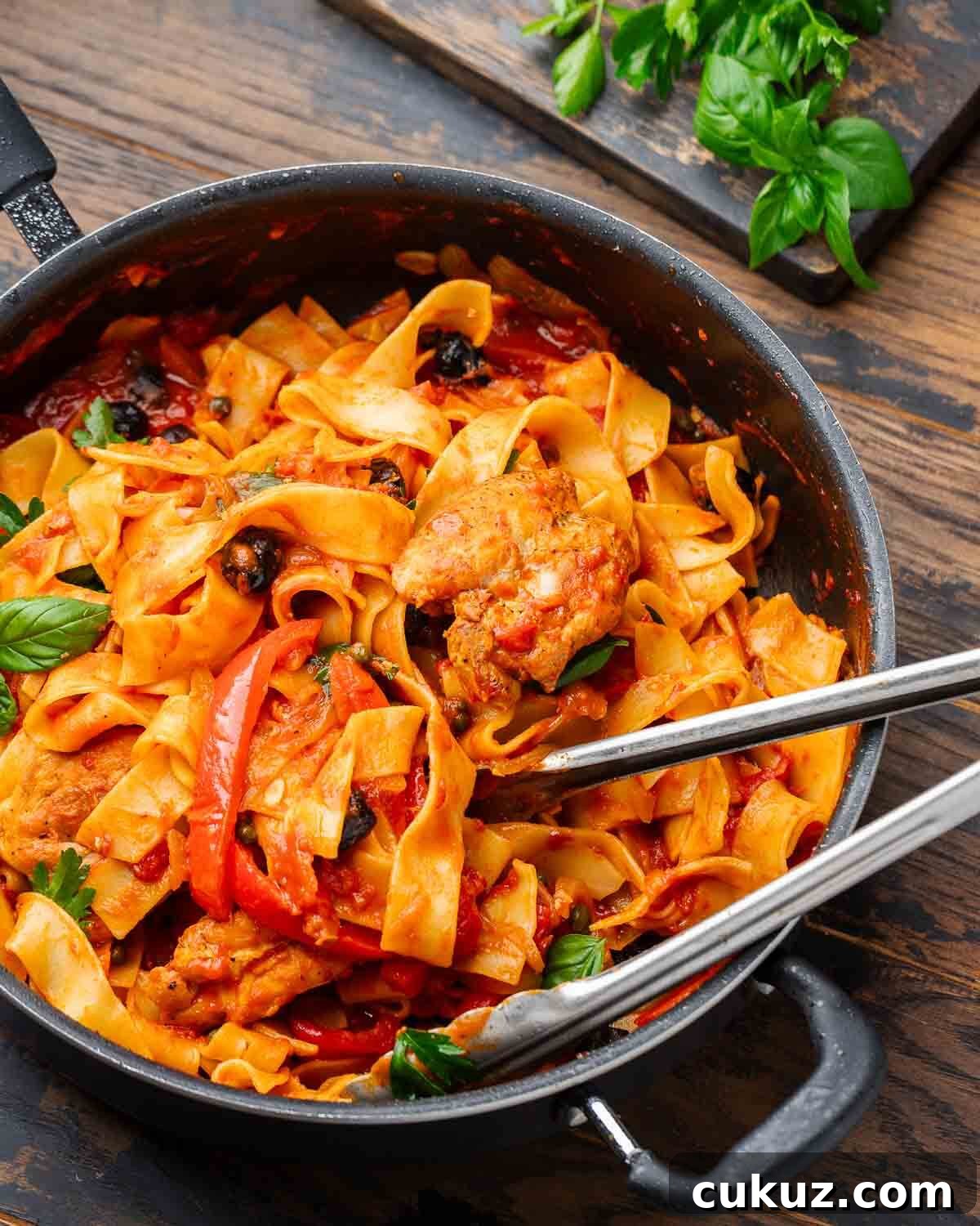 Large pan with chicken cacciatore pasta with tongs and board with basil and parsley.