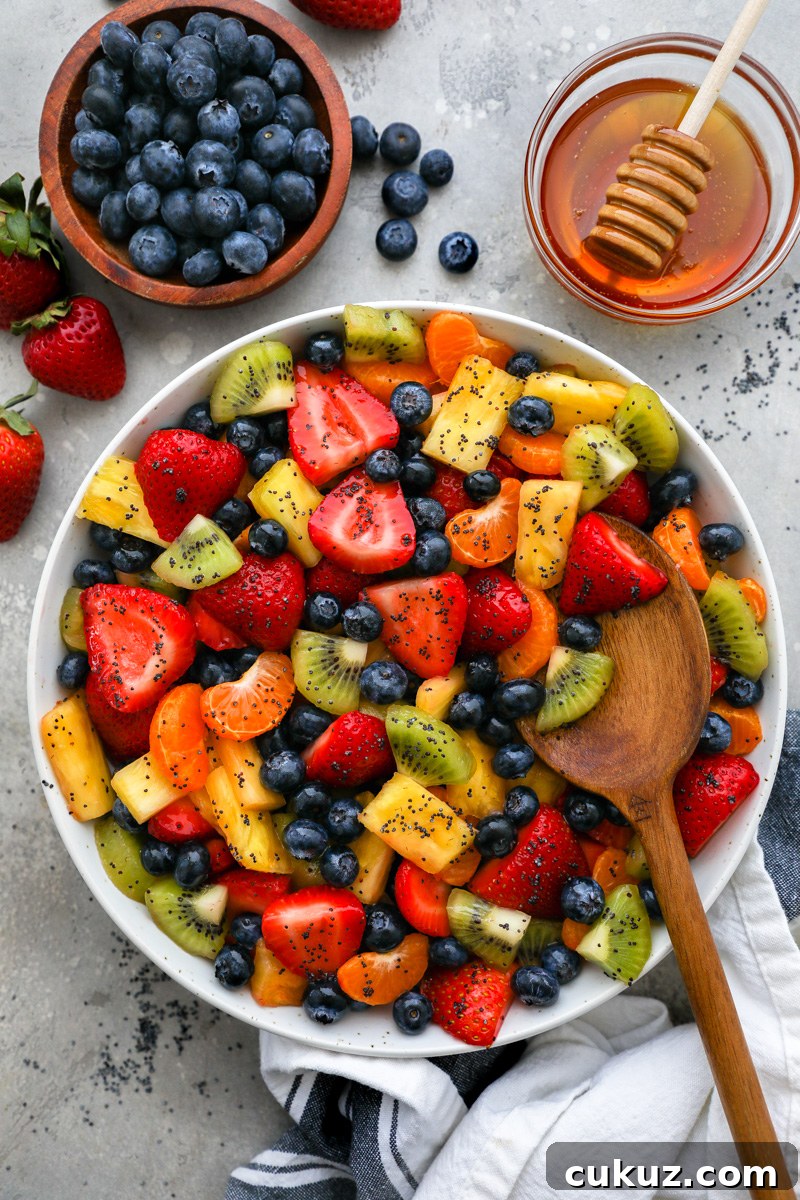 A large fresh fruit salad tossed in a honey lime poppy seed dressing surrounded by a bowl of blueberries, fresh strawberries and a jar filled with honey