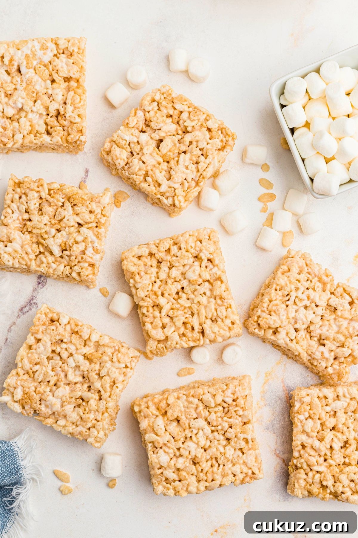 Rice krispie treats cut into squares on a marble table with mini marshmallows in a small bowl. 