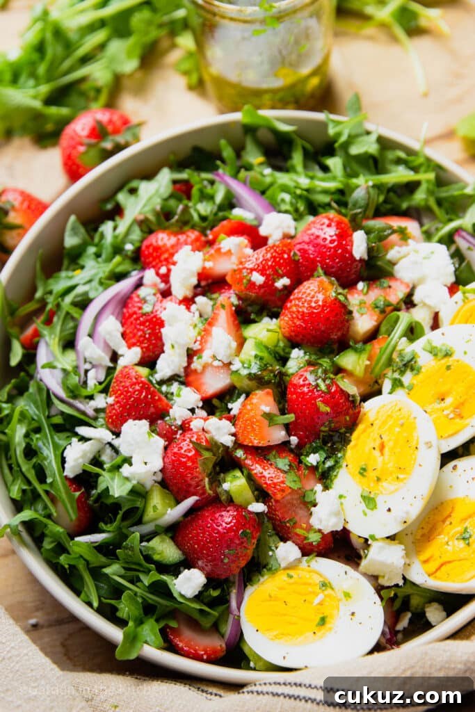 Salad in a bowl with strawberry, arugula, feta, red onions and hard boiled eggs. A jar of salad dressing and fresh strawberries on the table.