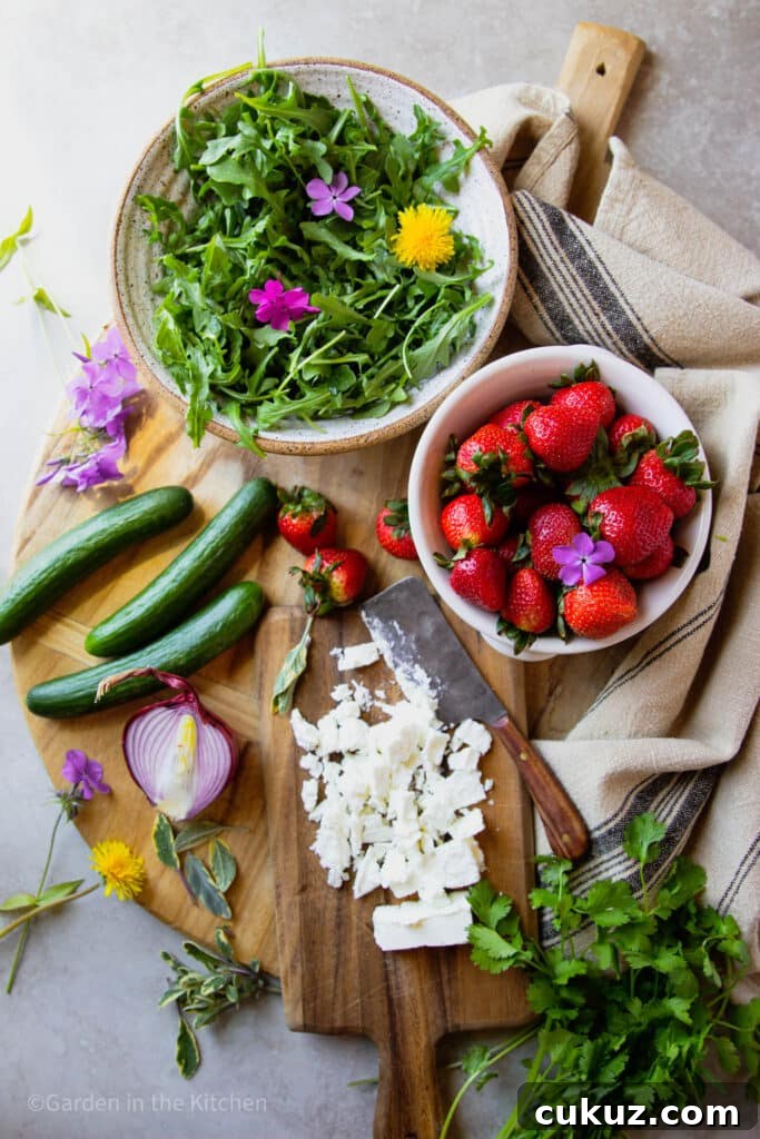 A big bowl with arugula and flowers. A smaller bowl with strawberries. Cucumbers, onion and feta cheese on a cutting board.