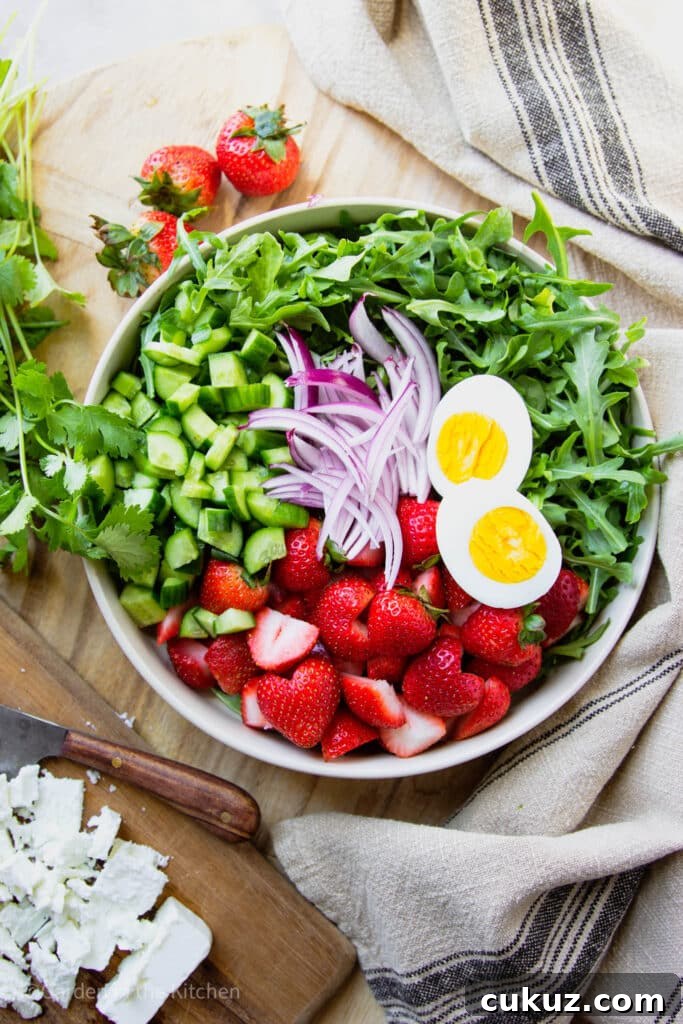 Chopped ingredients for a salad. Cucumbers, sliced strawberries, sliced red onions and hard boiled egg halved in a big bowl with arugula.