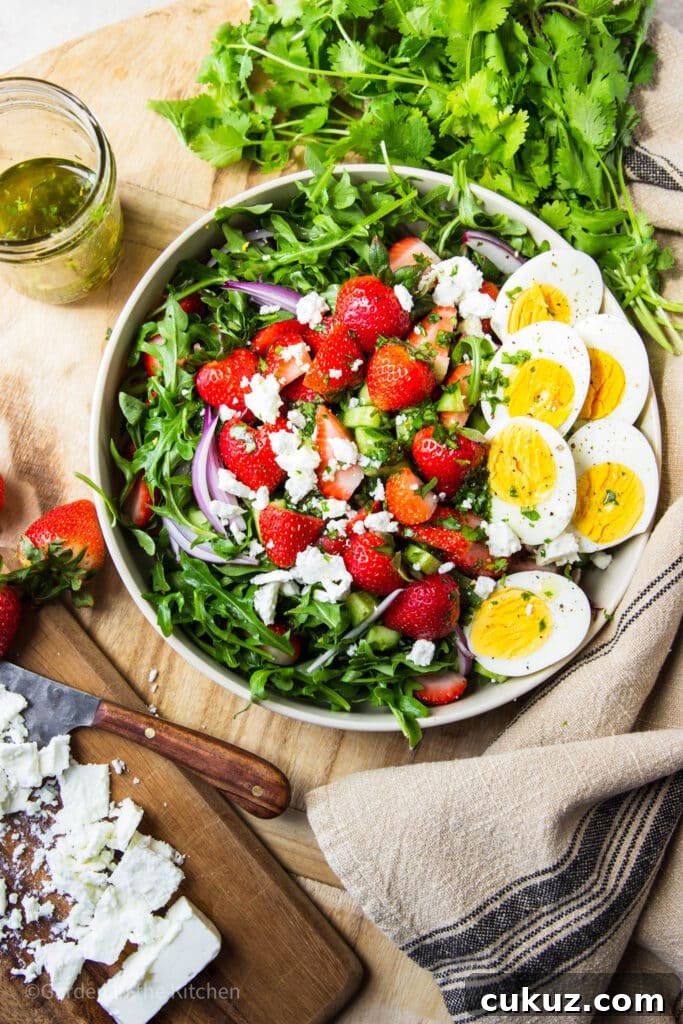 Salad in a bowl with strawberry, arugula, feta, red onions and hard boiled eggs. Fresh cilantro, a jar of dressing, strawberries and feta cheese on the table.