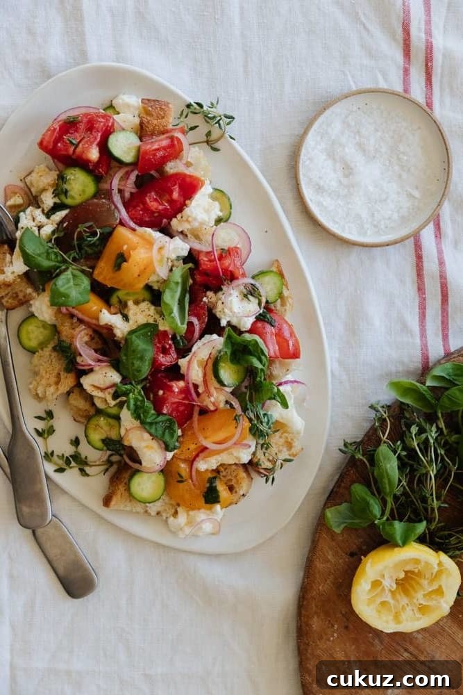 Rustic Panzanella Salad with Heirloom Tomatoes and Basil 3 panzanella salad on a white oval platter on top on a linen tablecloth with a salt pinch pot in the top right and fresh herbs and lemon in the bottom right.