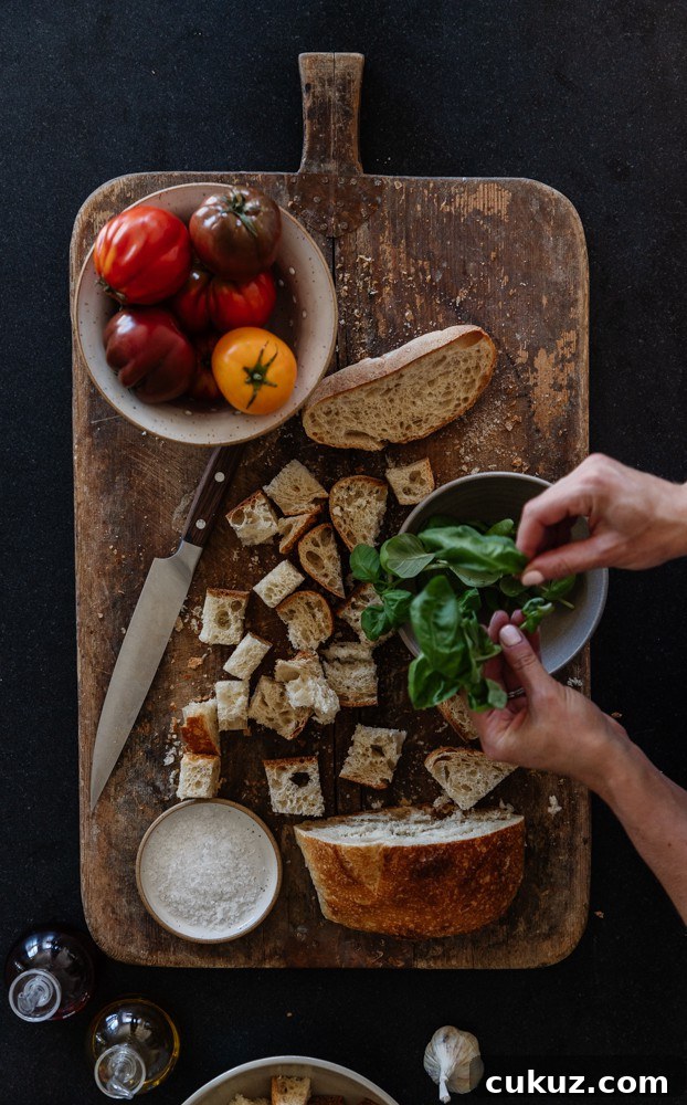 Rustic Panzanella Salad with Heirloom Tomatoes and Basil 4 rustic cutting board on black granite with cubed sourdough, bowl of tomatoes, and hands tearing some fresh basil