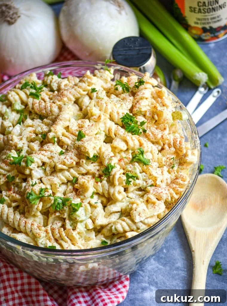creamy macaroni salad in a glass bowl on top of a red checkered cloth napkin with a wooden spoon and vegetables in the background