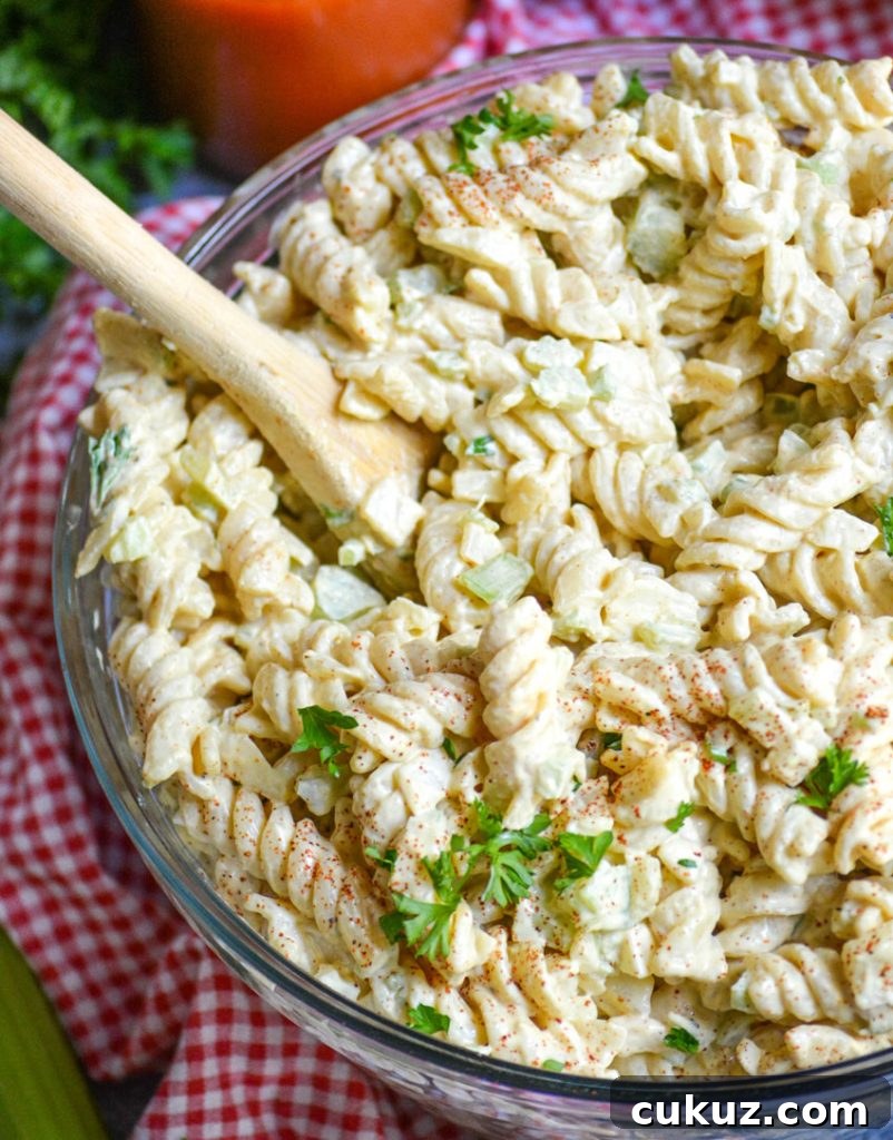 creamy macaroni salad in a glass bowl on top of a red checkered cloth napkin and a wooden spoon taking a scoop out