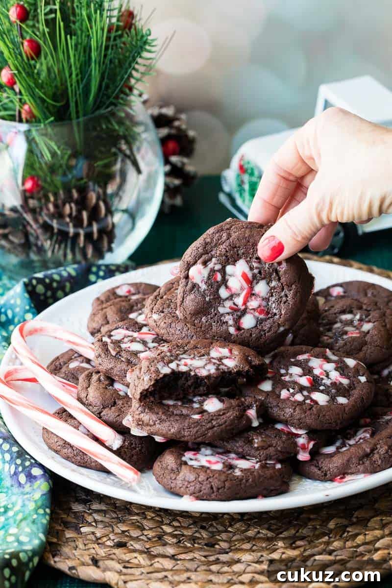 A hand lifts a peppermint chocolate cookie topped with crushed peppermint from a plate stacked with similar cookies; holiday decor and candy canes are nearby.