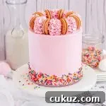 A pink Funfetti cake with brightly colored sprinkles all around the bottom of the cake on a white cake stand. The cake is topped with gold oreo cookies and pink frosting swirls.