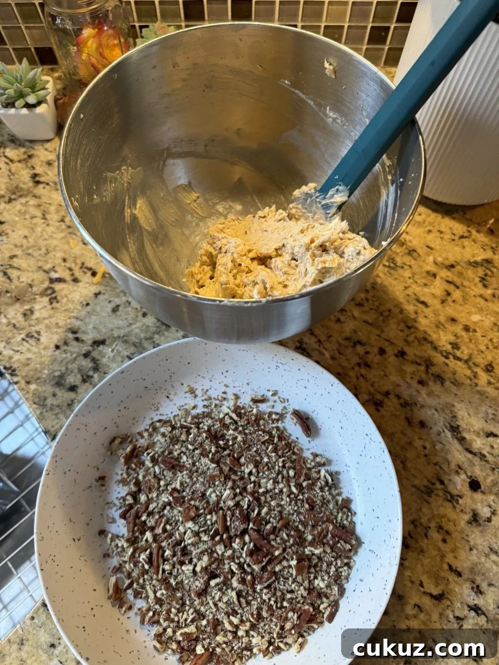 Bite-Sized Cheesy Snack Balls: Party Appetizer Recipe 5 The cheese ball mixture in a metal bowl with a blue spatula held over a white bowl of crushed pecans.