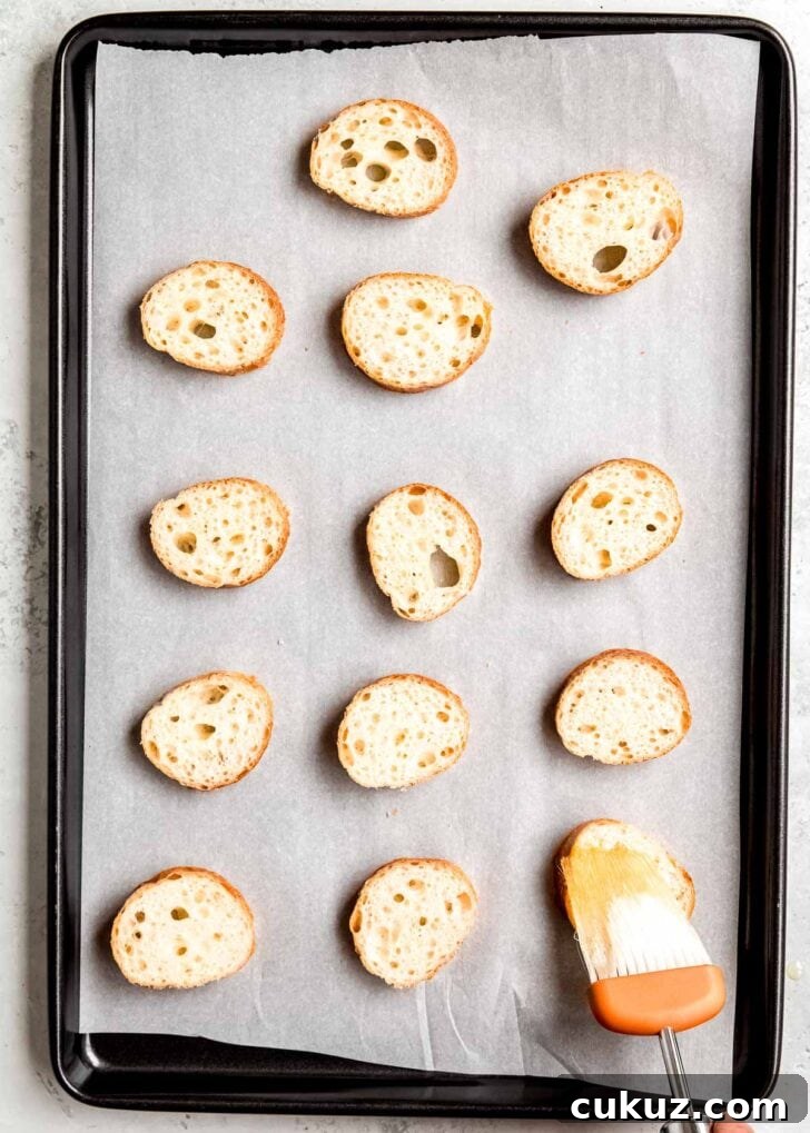 slices of french baguette on a parchment paper lined baking sheet. a culinary brush applying avocado oil to eat slice
