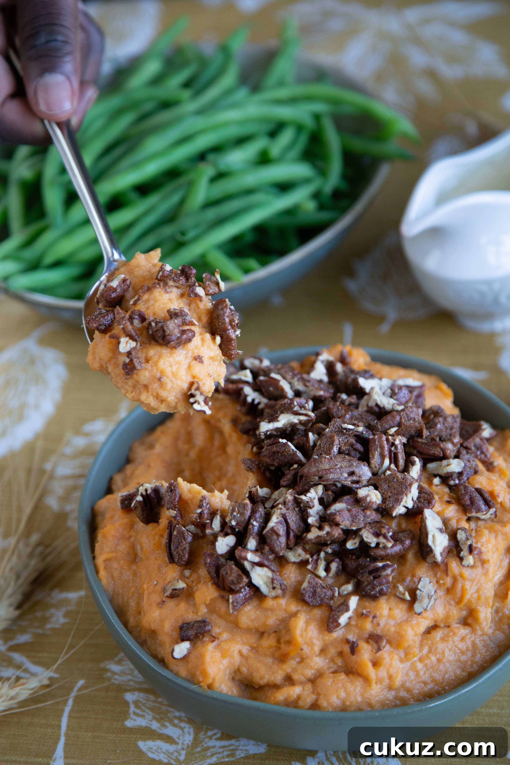 Mashed Sweet Potatoes with Cinnamon-Spiced Pecans 3 hand holding a spoonful of mashed sweet potatoes topped with spiced pecans on a table with fresh steamed green beans.