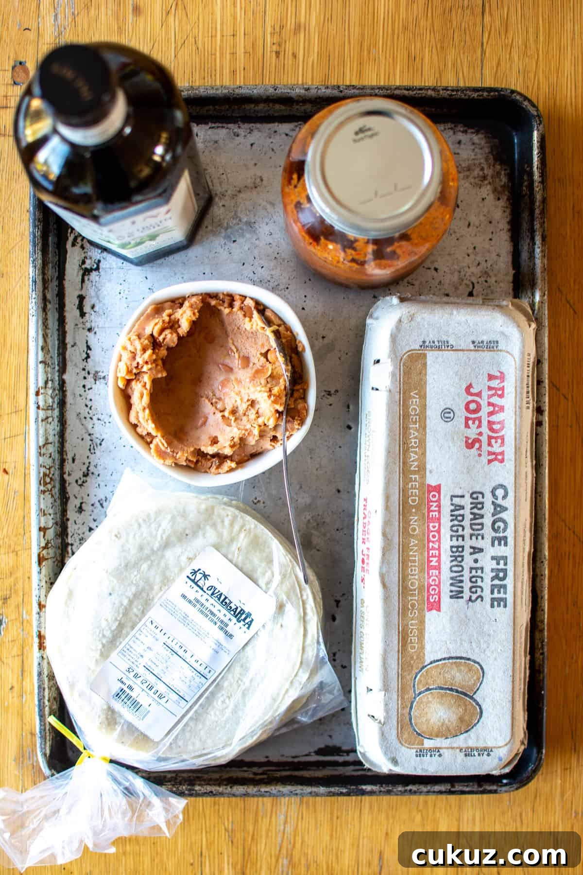 All the ingredients needed to make Huevos Rancheros including tortillas, eggs, beans, salsa, and olive oil on a metal baking dish. 