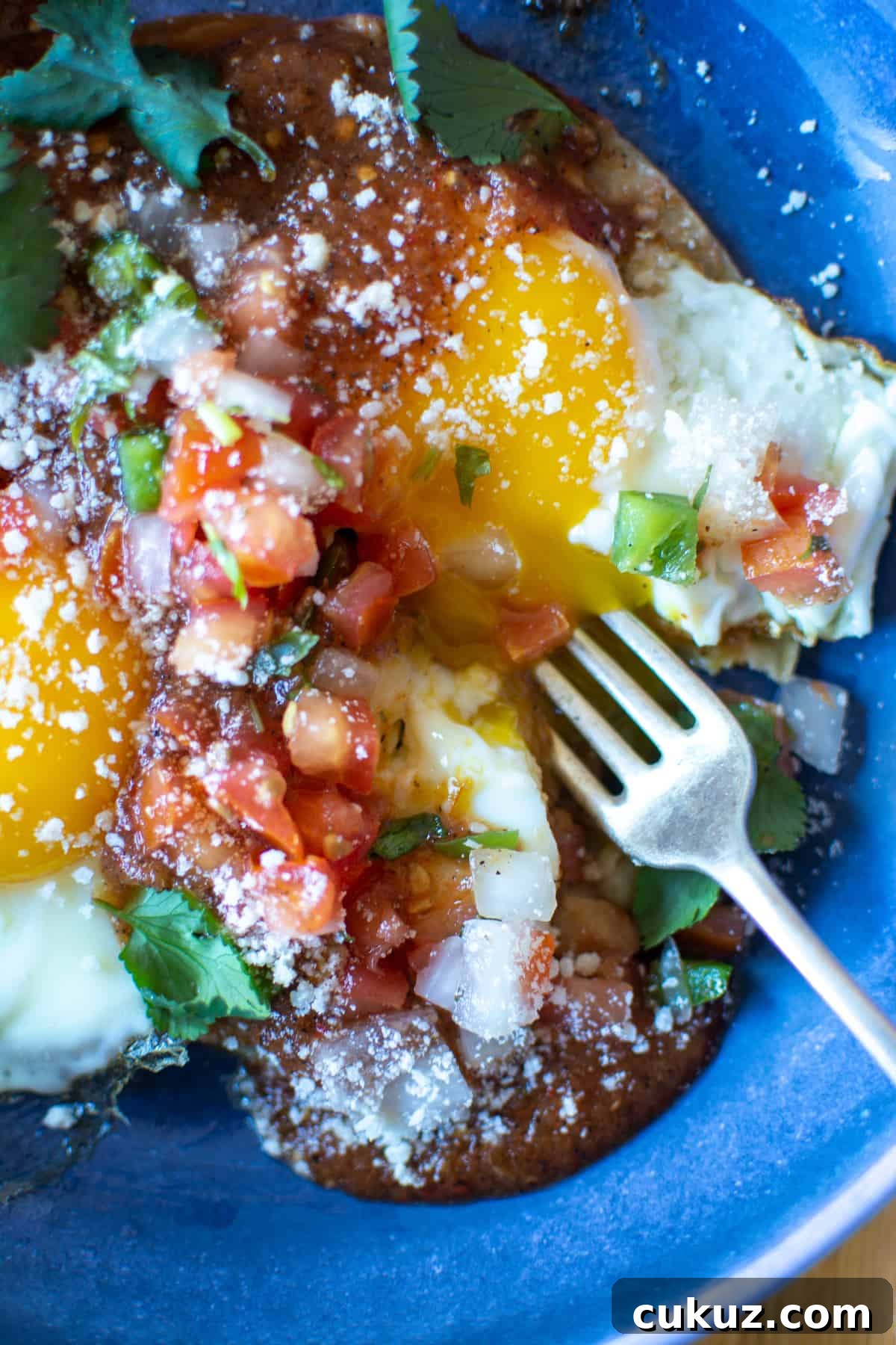 Fried eggs topped with Pico de Gallo and a fork breaking open the egg yolk in a blue plate with Cotija cheese on top. 